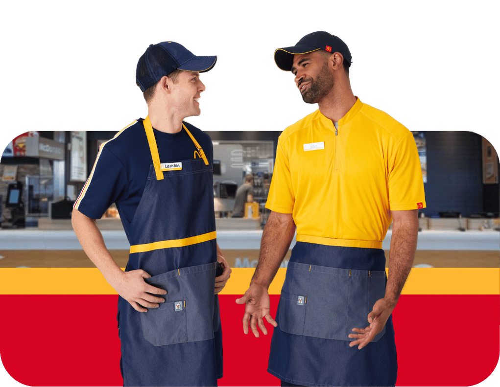 Two McDonald's employees wearing yellow and navy McDonald’s uniforms and aprons, standing together inside a restaurant. Two McDonald's employees wearing yellow and navy McDonald’s uniforms and aprons, standing together inside a restaurant.